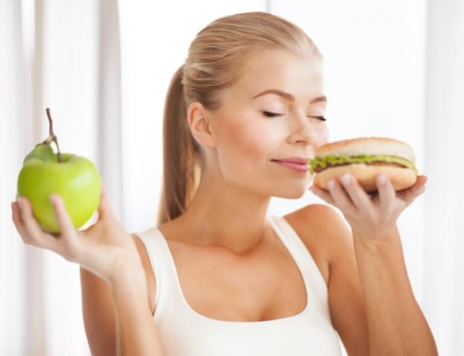 woman-in-white-shirt-smelling-burger-while-holding-apple